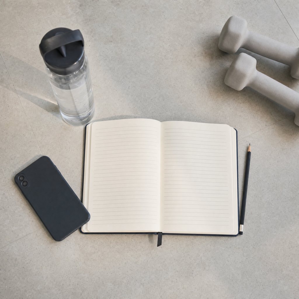 An open training notebook with a pencil beside a phone, a water bottle and a pair of light dumbbells on a pale floor